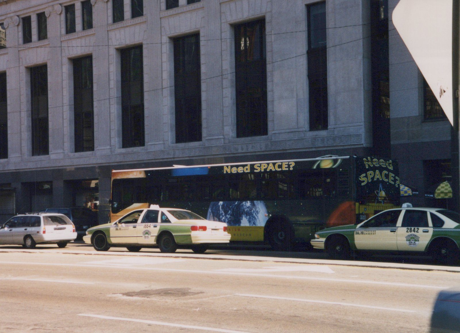 A side view of a Chicago Transit Authority bus with a large 'Need SPACE?' ad on its side, driving down a street with green and white taxis nearby.