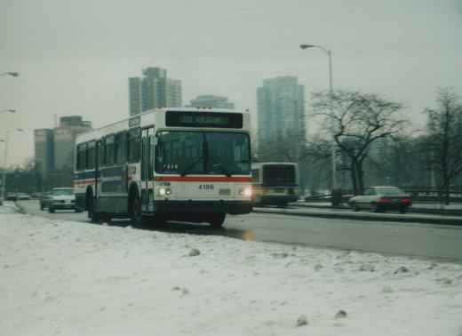 A white CTA bus with red and blue stripes, numbered 4196, drives on a snowy city street with tall buildings in the background.