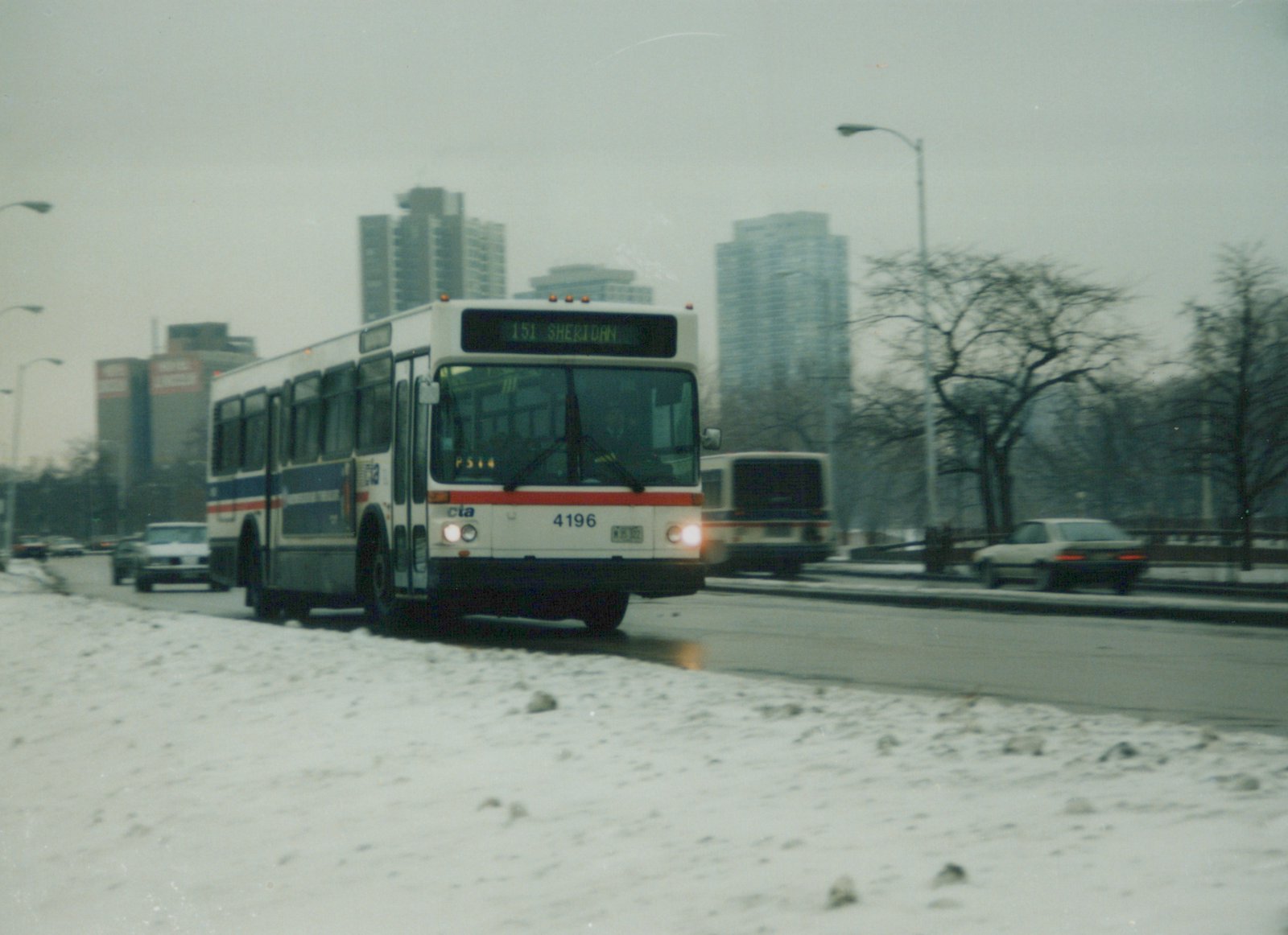 A white CTA bus with red and blue stripes, numbered 4196, drives on a snowy city street with tall buildings in the background.