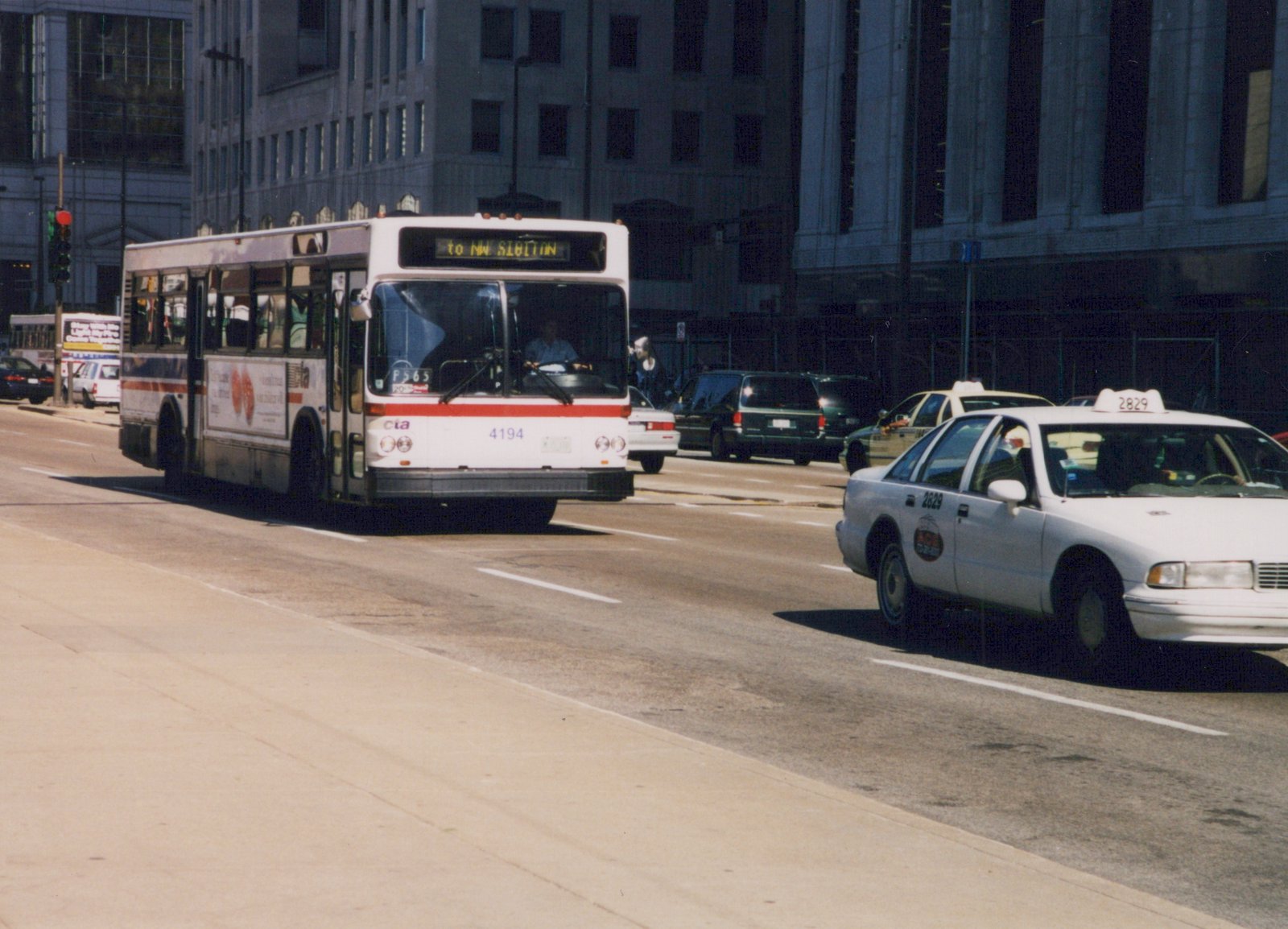 A white CTA bus with red stripe and number 4194 drives down a city street alongside a white taxi, with large buildings in the background.