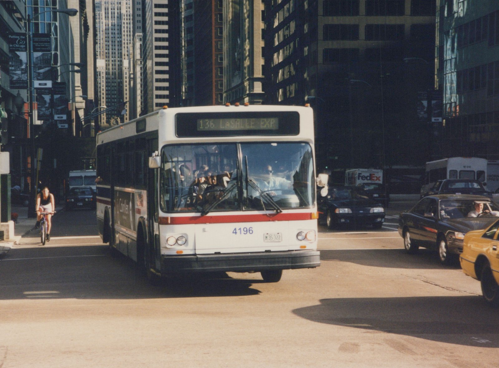 A white and red city bus with the number 4196 and destination sign reading 136 LaSalle Exp is driving on a city street with tall buildings, other cars, and a cyclist on the sidewalk.