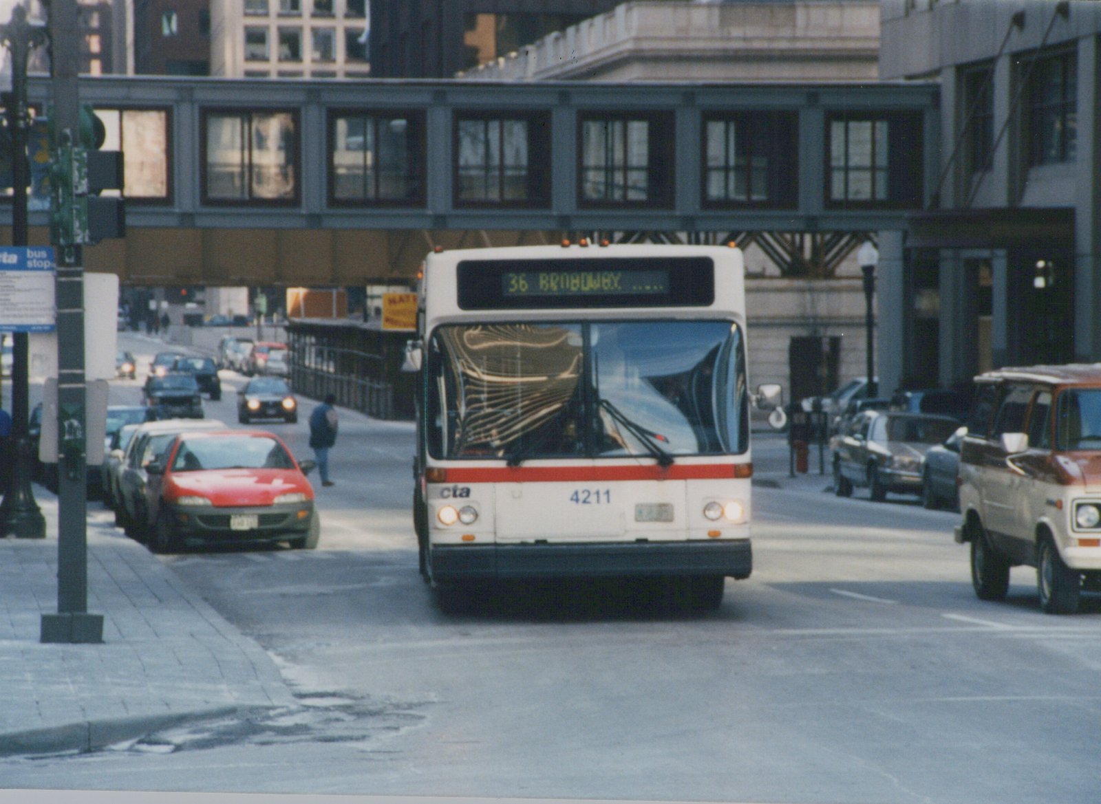 A white and red CTA bus, number 4211, travels down a city street under an elevated structure, with other cars and a pedestrian visible.