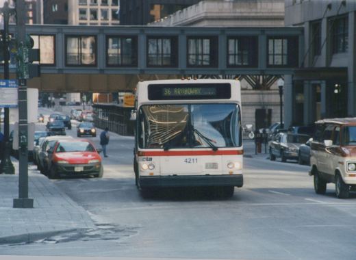 A white and red CTA bus, number 4211, travels down a city street under an elevated structure, with other cars and a pedestrian visible.