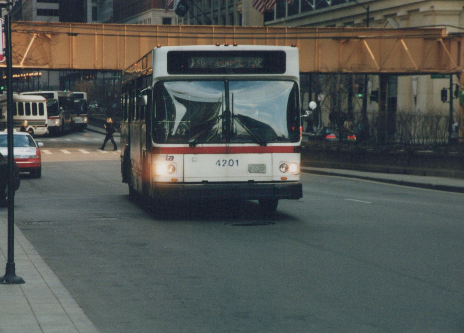 A white and red CTA bus, number 4201, is driving on a city street with an elevated railway track above it, and other vehicles and pedestrians are visible.
