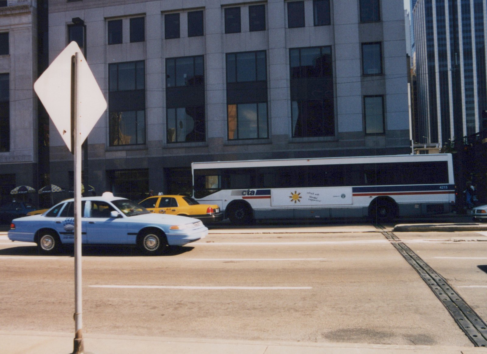A white and blue Chicago Transit Authority bus is on a city street with two taxis, one light blue and one yellow, in front of a modern building.