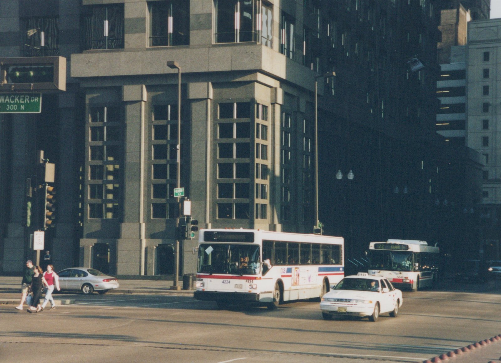 A white CTA bus with red and blue stripes, fleet number 4224, turns at an intersection with tall buildings and a green Wacker Drive street sign, while pedestrians cross and other buses and cars are nearby.