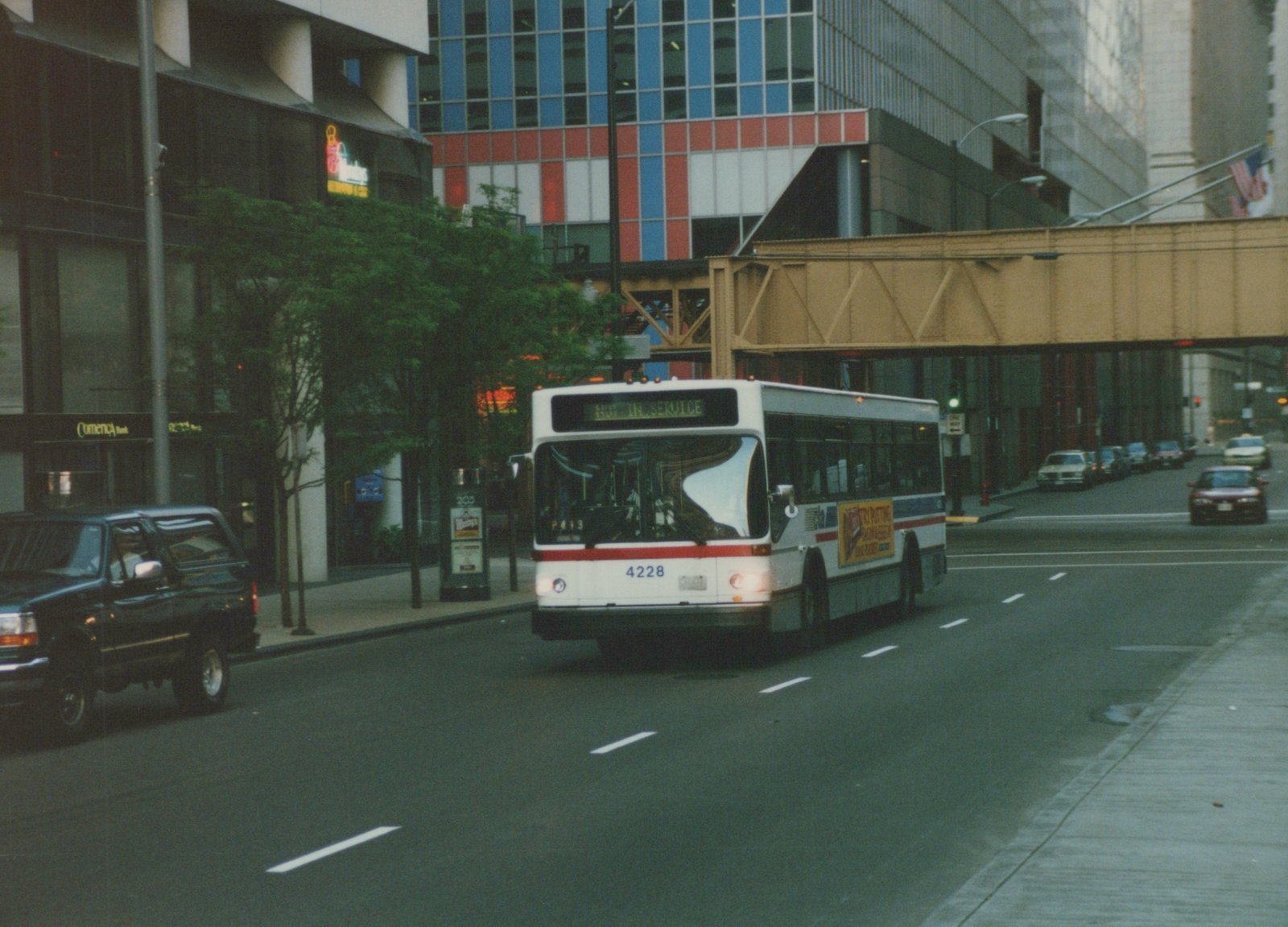A white and red CTA bus numbered 4228 drives down a city street under an elevated train bridge, with other cars and buildings visible.