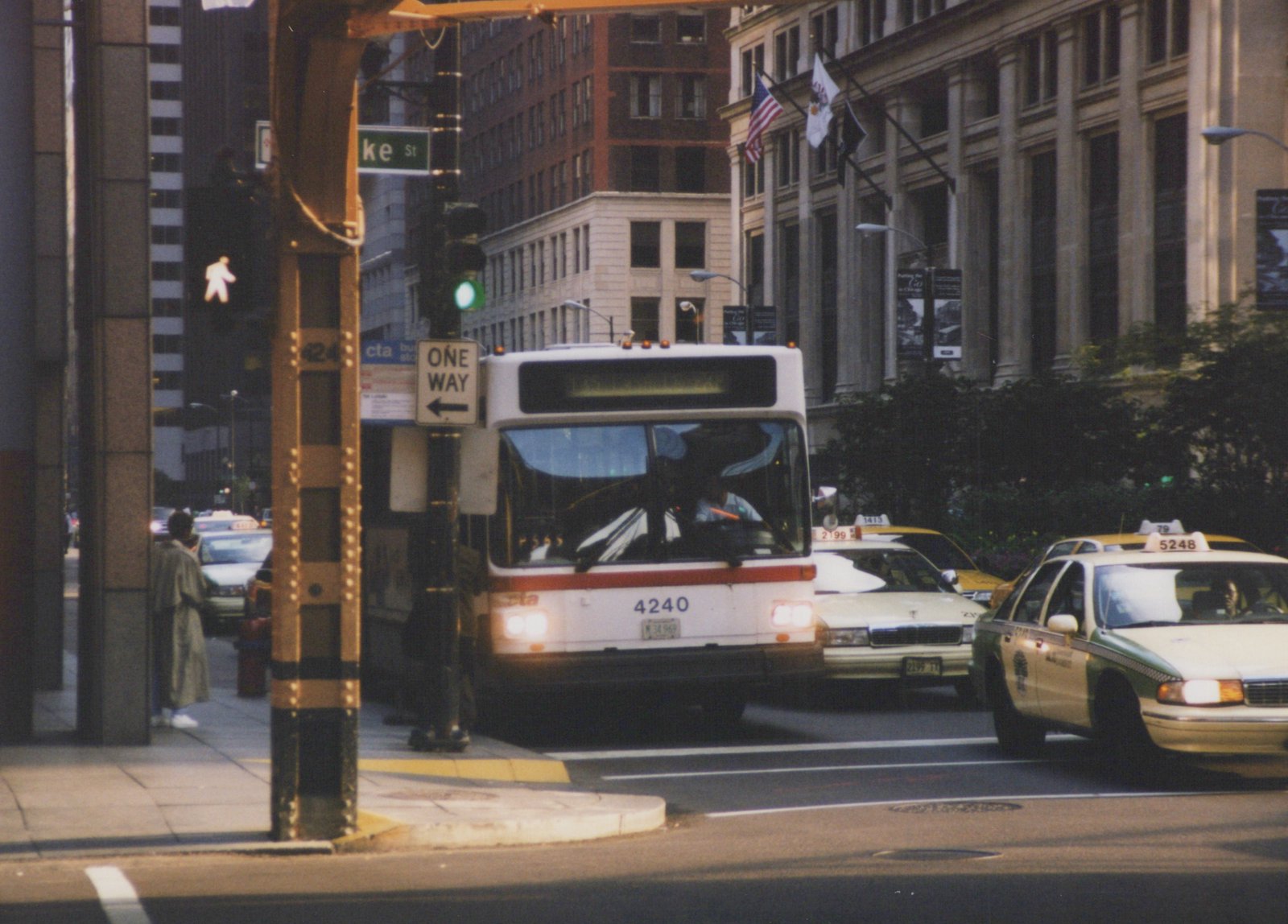 A white CTA bus with the number 4240 on the front is driving on a city street. There are several taxicabs nearby and tall buildings in the background.