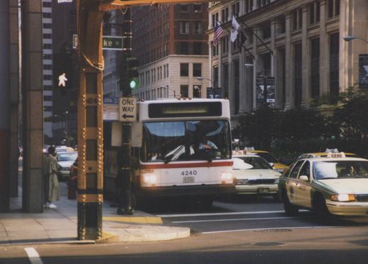 A white CTA bus with the number 4240 on the front is driving on a city street. There are several taxicabs nearby and tall buildings in the background.