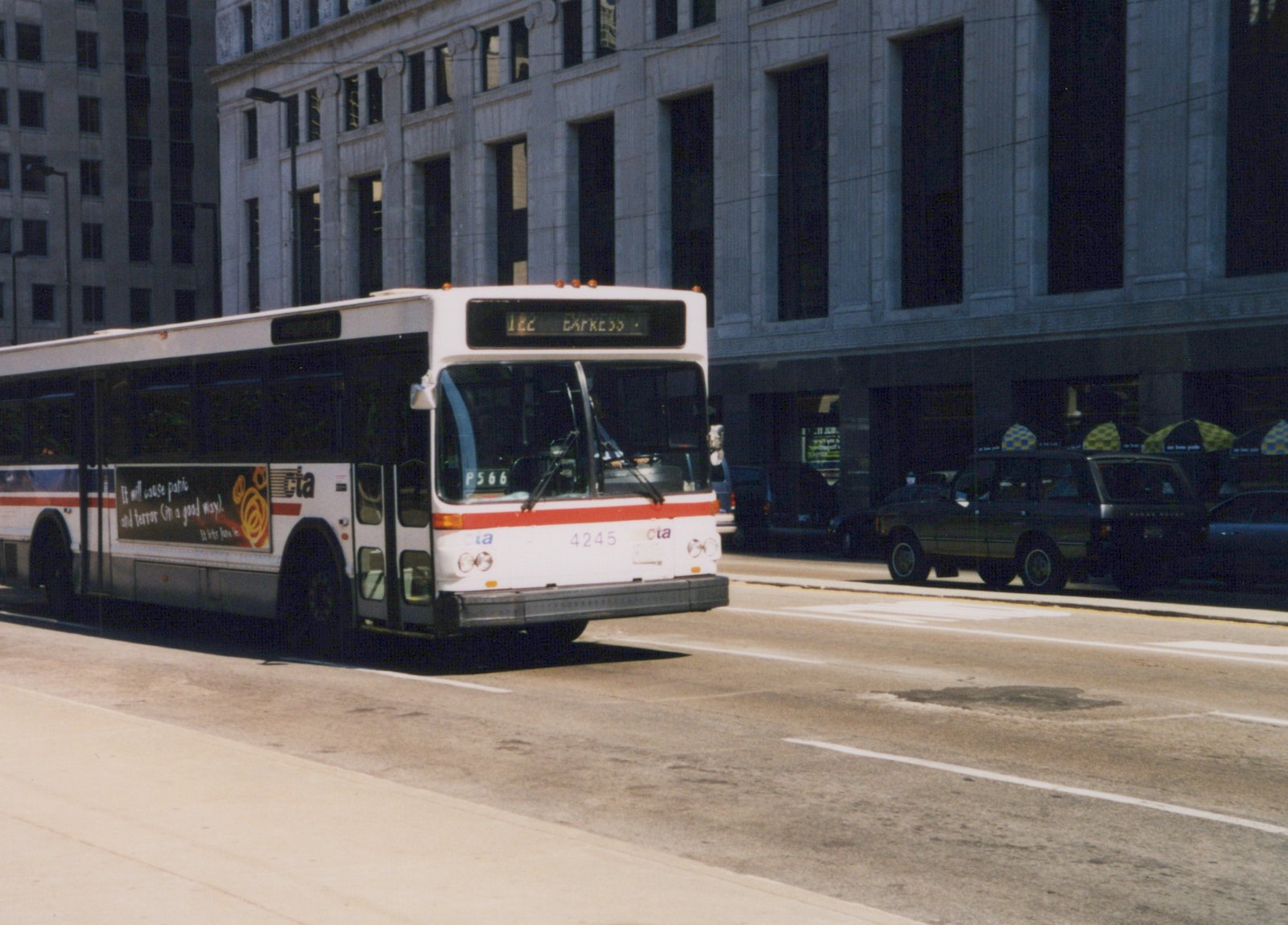 A white and red CTA bus with the number 4245 is driving down a city street, with a large stone building in the background and other vehicles parked nearby.