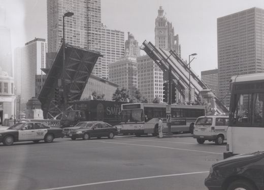 A black and white photo of a CTA bus at a street intersection with a large, raised drawbridge in the background and tall buildings behind it.