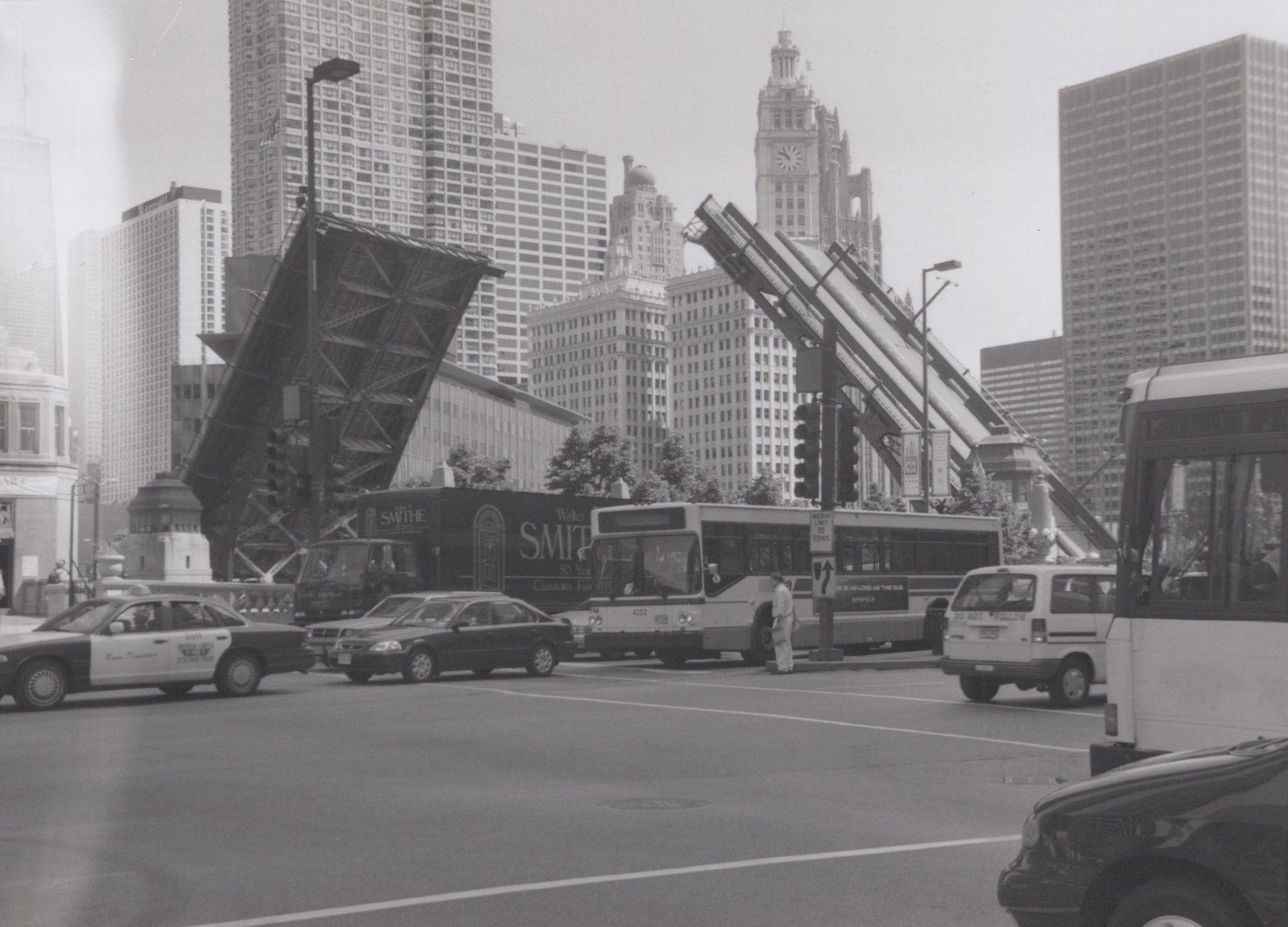 A black and white photo of a CTA bus at a street intersection with a large, raised drawbridge in the background and tall buildings behind it.