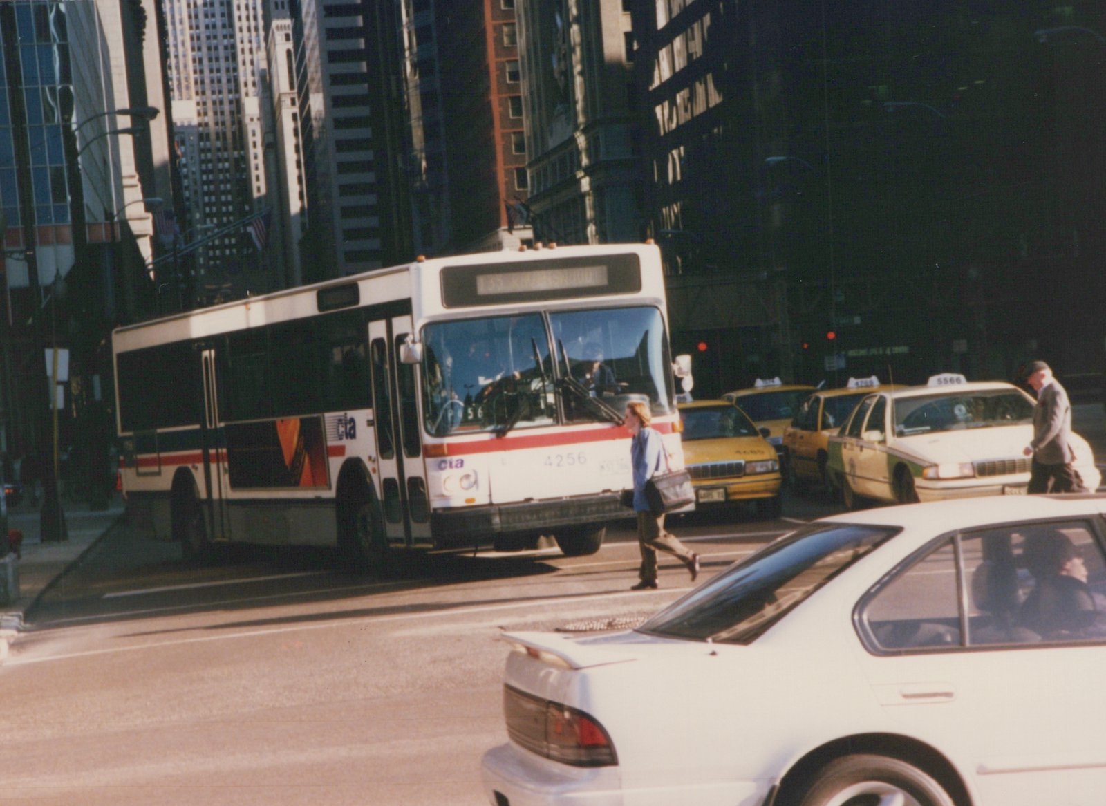 A white CTA bus with the number 4256 on its front is stopped on a city street, surrounded by yellow taxis and pedestrians in an urban setting with tall buildings in the background.