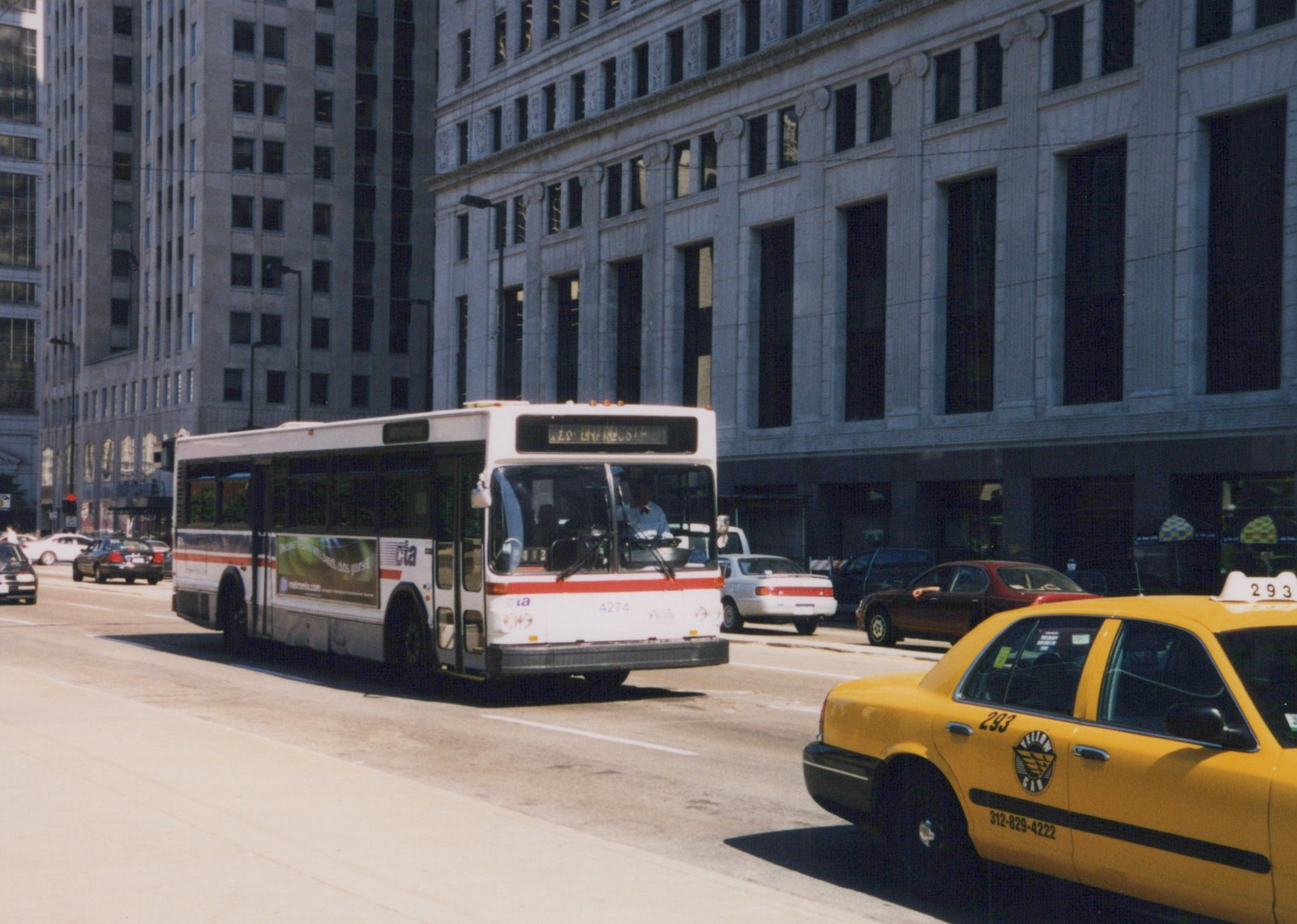 A white and red Chicago Transit Authority bus drives down a city street next to a yellow taxi, with tall buildings in the background.