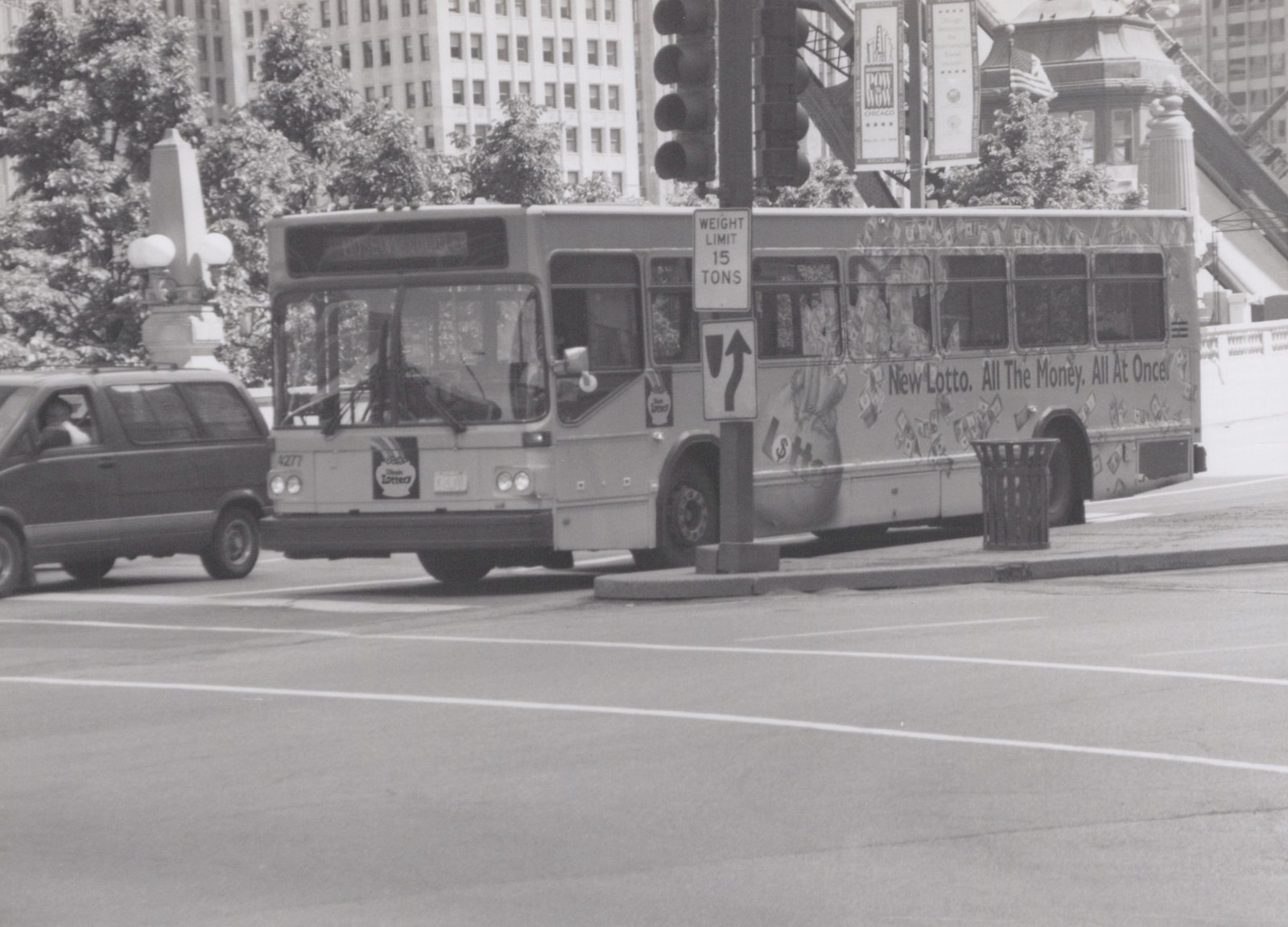 A CTA bus with the number 4277 and an Illinois Lottery ad is stopped on a city street, with a minivan beside it and banners in the background.