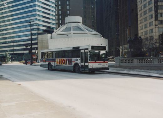 A white and blue Chicago Transit Authority bus, number 4283, drives down a snow-covered street with tall buildings and a modern church in the background.