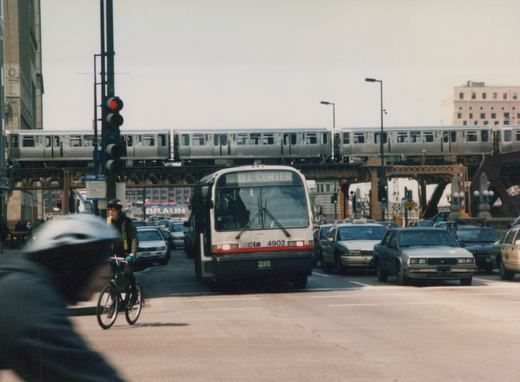 A white CTA bus with the number 4902 is driving down a street with a silver CTA train on an elevated track above. A bicyclist and several cars are also on the street.