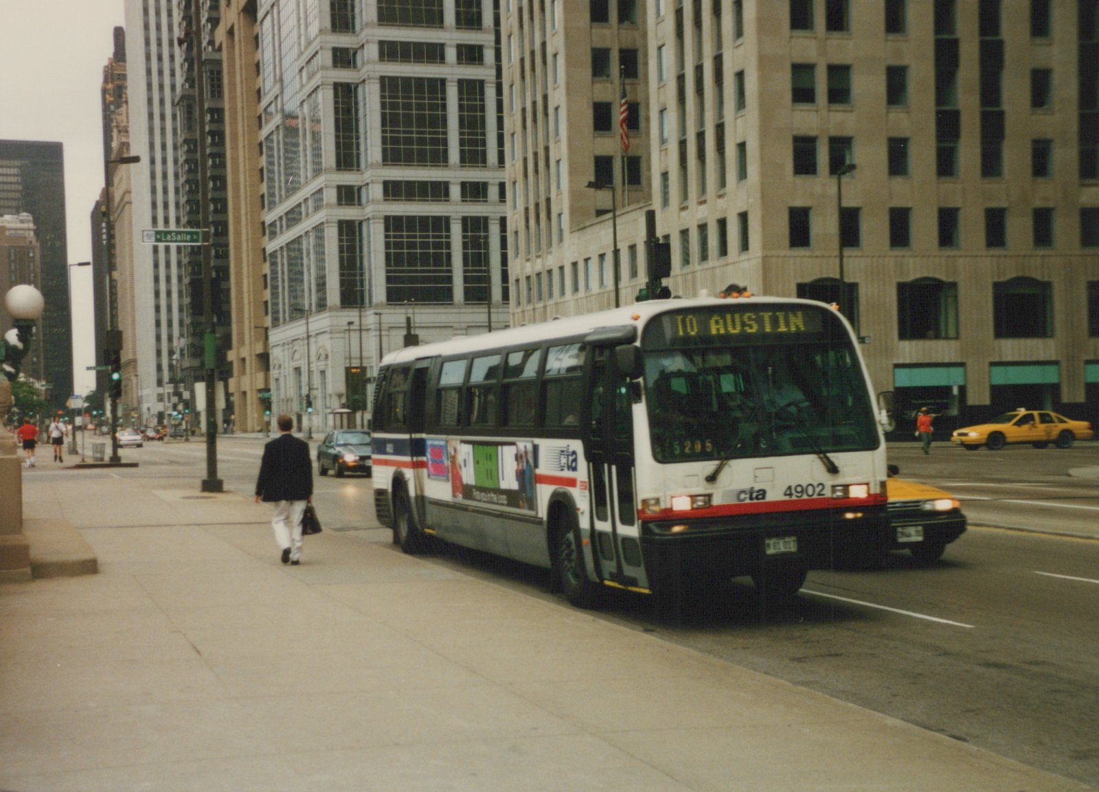 A white CTA bus with the number 4902 and destination TO AUSTIN is driving down a city street lined with tall buildings, with pedestrians on the sidewalk.
