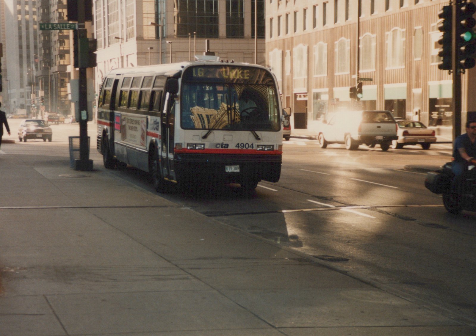 A white and red Chicago Transit Authority bus on a city street with a destination sign reading '16 LAKE', with buildings and other vehicles in the background.