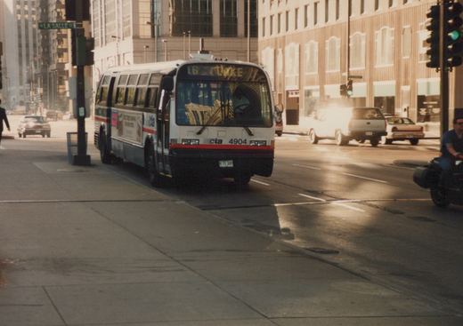 A white and red Chicago Transit Authority bus on a city street with a destination sign reading '16 LAKE', with buildings and other vehicles in the background.