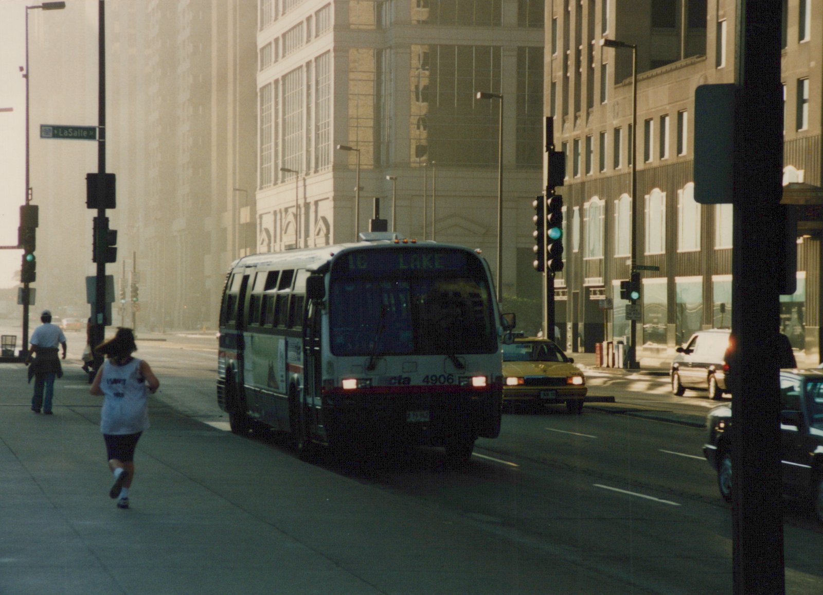 A white and blue Chicago Transit Authority bus with the number 4906 on the front is turning at an intersection with a LaSalle Street sign visible; people are walking and jogging on the sidewalk.