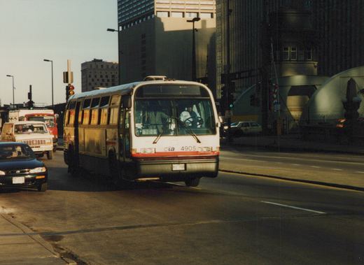 A white and red CTA bus, fleet number 4905, drives on a city street with other cars and tall buildings in the background on a clear day.