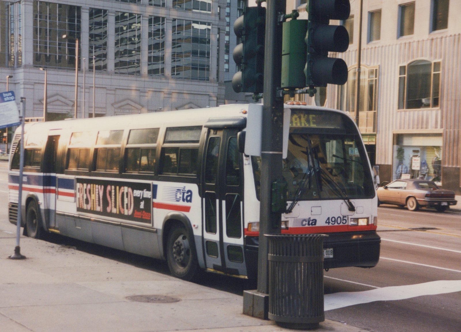 A white Chicago Transit Authority bus with the number 4905 is parked on a city street next to a traffic light and trash can, with a tan car in the background.