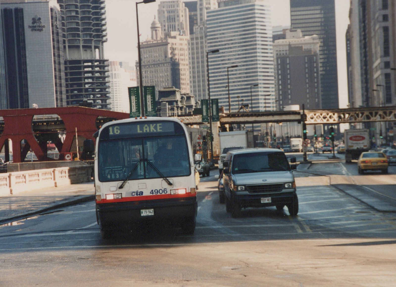 Front view of a white Chicago Transit Authority bus with the number 4906 and route 16 Lake displayed, driving on a city street with tall buildings and a red bridge in the background.