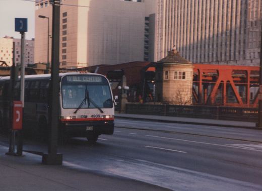 A white and red CTA bus, number 4909, is driving on a city street in front of a modern building and a red bridge structure.