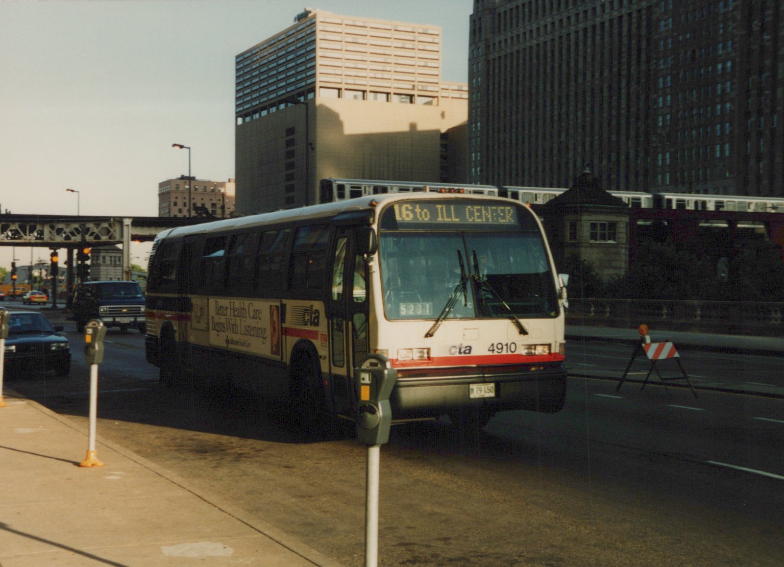 A white CTA bus with red stripes, number 4910, drives down a Chicago street with an elevated train and tall buildings in the background.