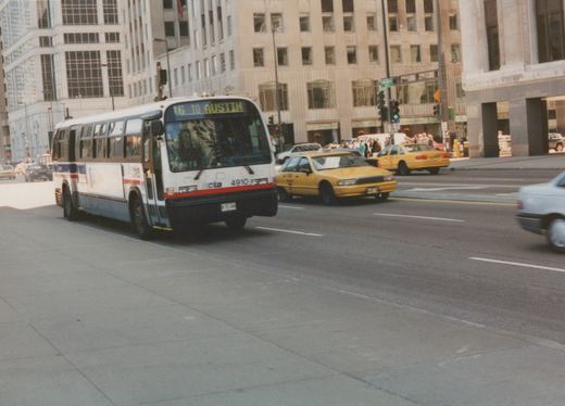A white, blue, and red CTA bus with the number 4910 is driving down a city street with yellow taxis and tall buildings in the background.