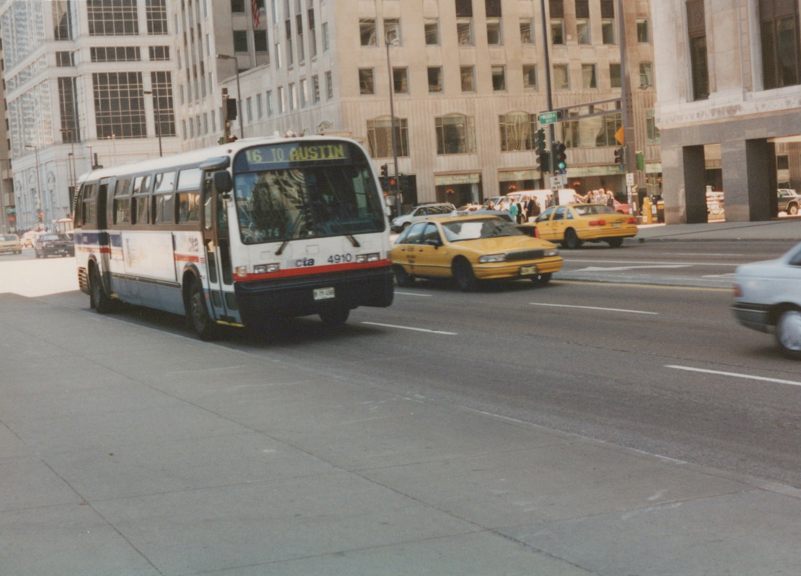 A white, blue, and red CTA bus with the number 4910 is driving down a city street with yellow taxis and tall buildings in the background.