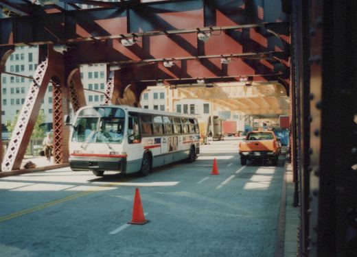 A white and red Chicago Transit Authority bus numbered 4913 is driving on a bridge under a red truss structure, with an orange pickup truck visible behind it.