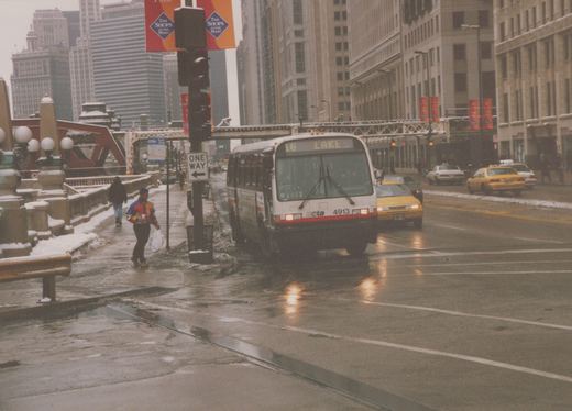 A white and red CTA bus with the number 4913 and destination '16 LAKE' is stopped on a wet, snow-lined city street with pedestrians and yellow taxis nearby.