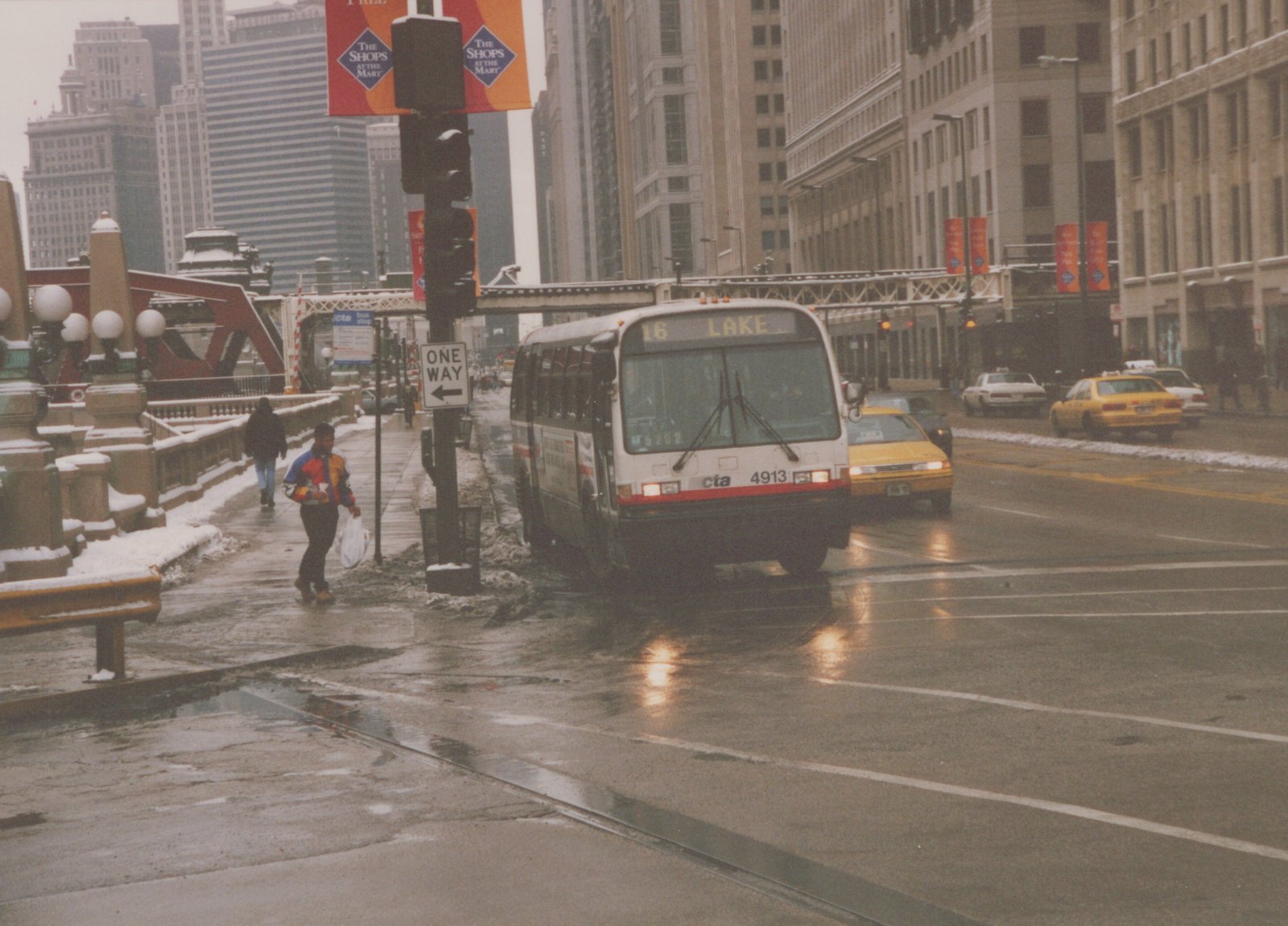 A white and red CTA bus with the number 4913 and destination '16 LAKE' is stopped on a wet, snow-lined city street with pedestrians and yellow taxis nearby.