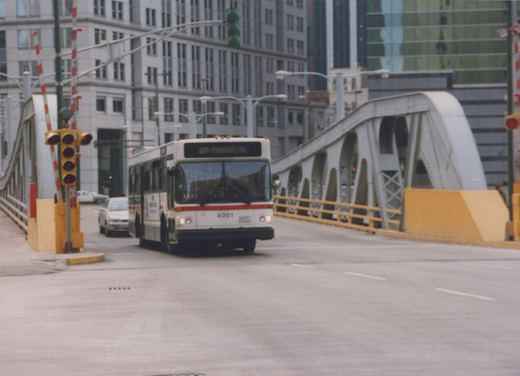 A white and red CTA bus, number 4001, is driving on a bridge in an urban setting with tall buildings in the background.
