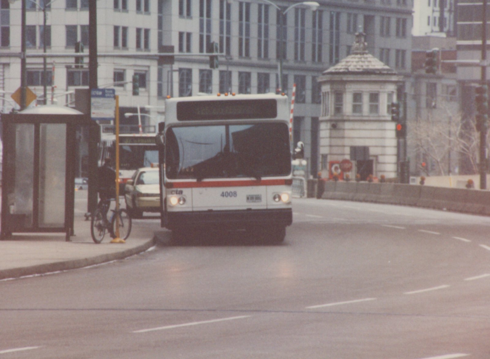 A white and orange Chicago Transit Authority bus with the number 4008 on the front is stopped on a city street next to a bus shelter, with a person standing with a bicycle nearby.