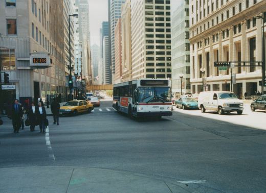 A white, red and blue CTA bus drives down a city street lined with tall buildings, with pedestrians crossing and other cars nearby.