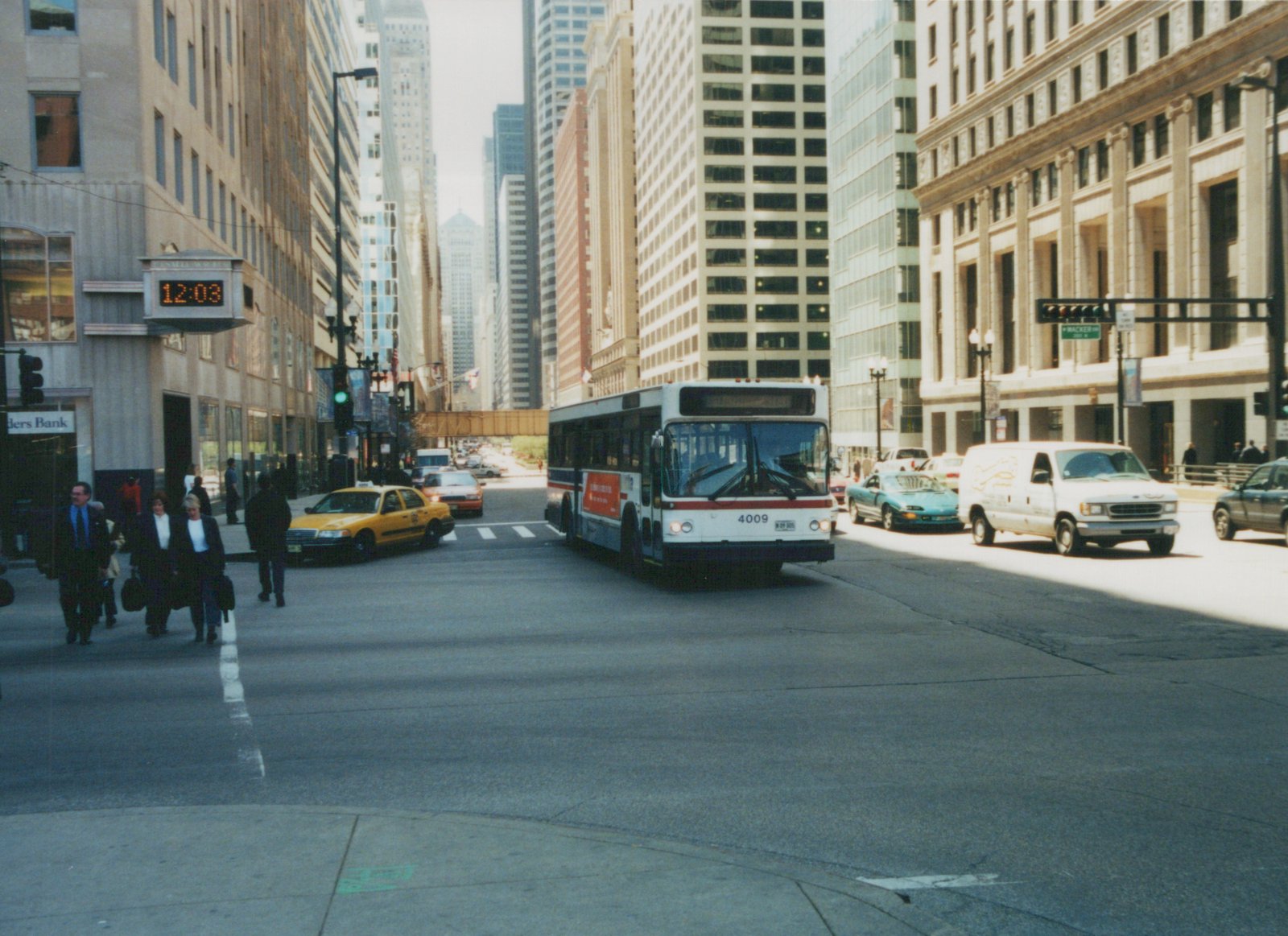 A white, red and blue CTA bus drives down a city street lined with tall buildings, with pedestrians crossing and other cars nearby.
