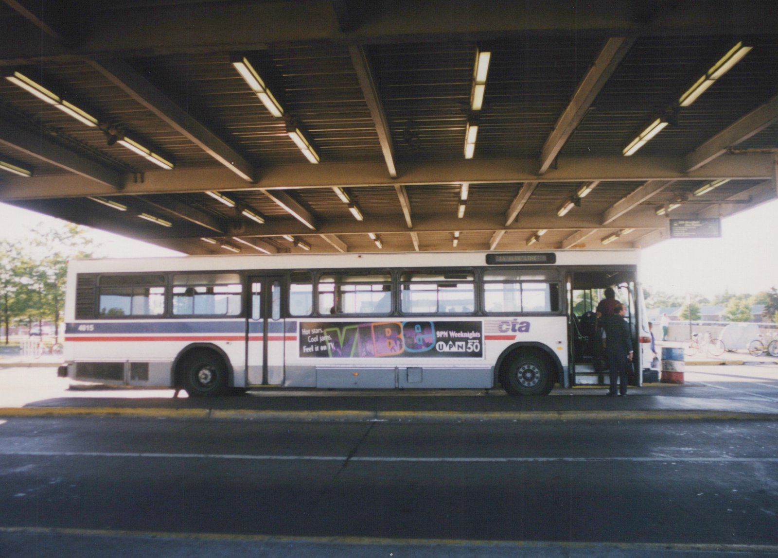 A white, blue, and red Chicago Transit Authority bus numbered 4015 is stopped at a covered terminal with passengers entering. The bus displays an ad for UPN 50 and is marked for Route 46 Blue Line.