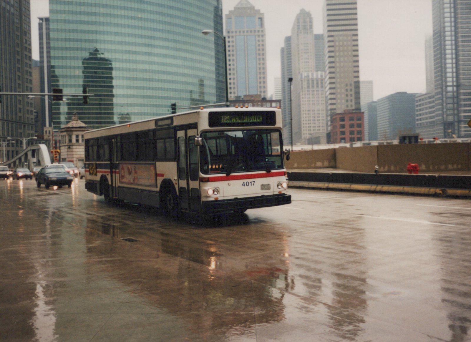 A white and red Chicago Transit Authority bus, number 4017, is driving on a wet street in front of tall glass buildings on a rainy day.