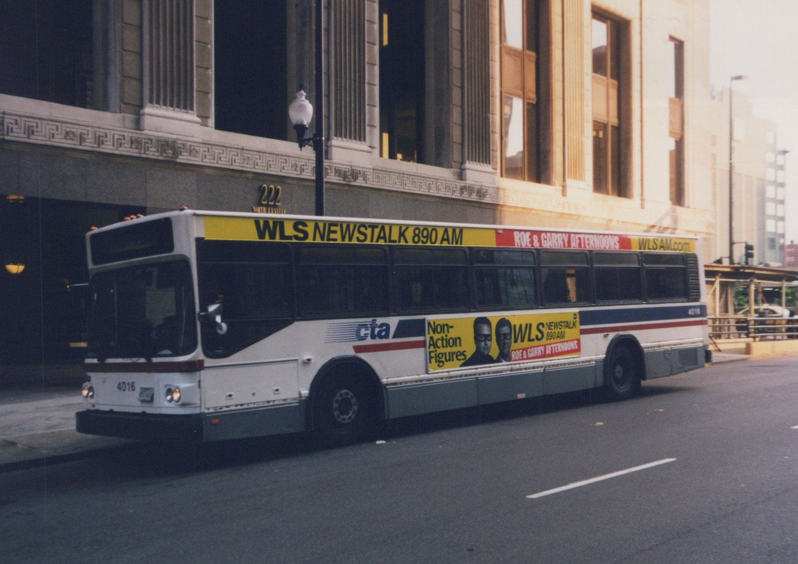 A white, blue, and red Chicago Transit Authority bus number 4016 is parked on a city street, with advertisements for WLS NewsTalk 890 AM on its side and roof, in front of a building marked 222 North LaSalle.