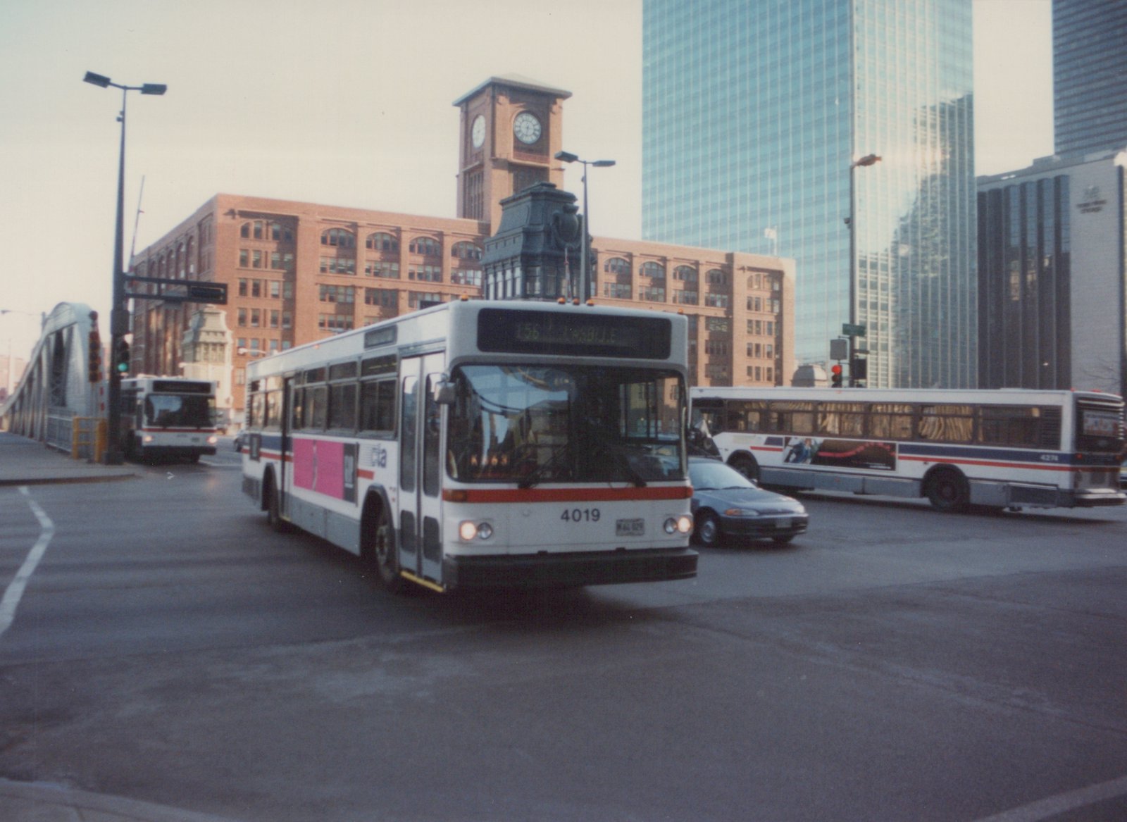 A white, red, and blue Chicago Transit Authority bus, number 4019, is turning on a city street with a clock tower and skyscrapers in the background.
