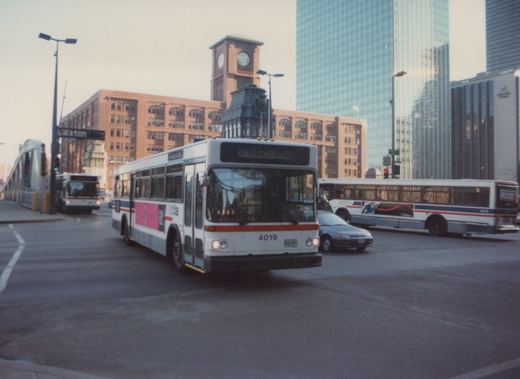 A white, red, and blue Chicago Transit Authority bus, number 4019, is turning on a city street with a clock tower and skyscrapers in the background.