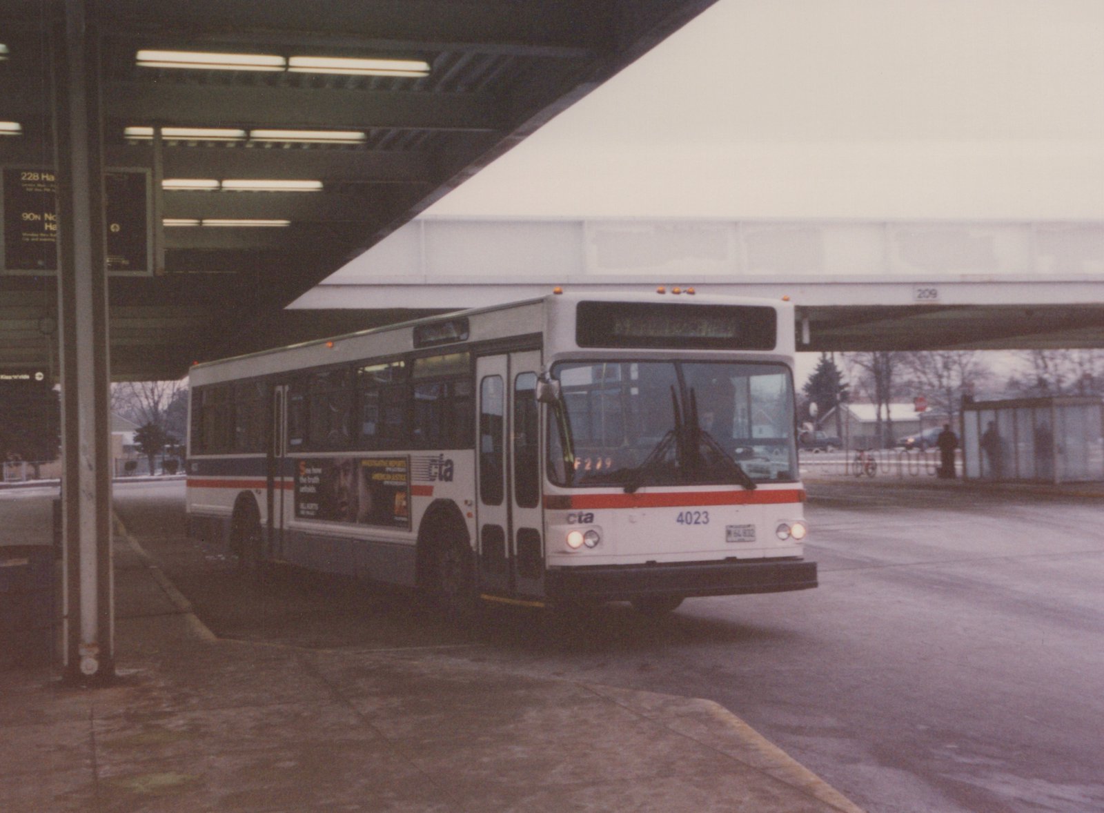 A white and red CTA bus numbered 4023 is parked under a concrete overpass at a bus stop. Two people are visible at a shelter in the background.
