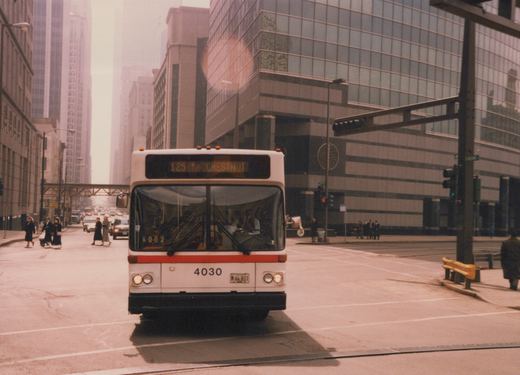 A white and red Chicago Transit Authority bus, number 4030, is stopped at a city intersection with tall buildings in the background and pedestrians nearby.