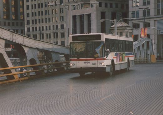 A white CTA bus, number 4030, is shown from a three-quarter front view driving on a street bridge in a city. A pedestrian walks on the bridge's walkway alongside the bus, with large buildings in the background.