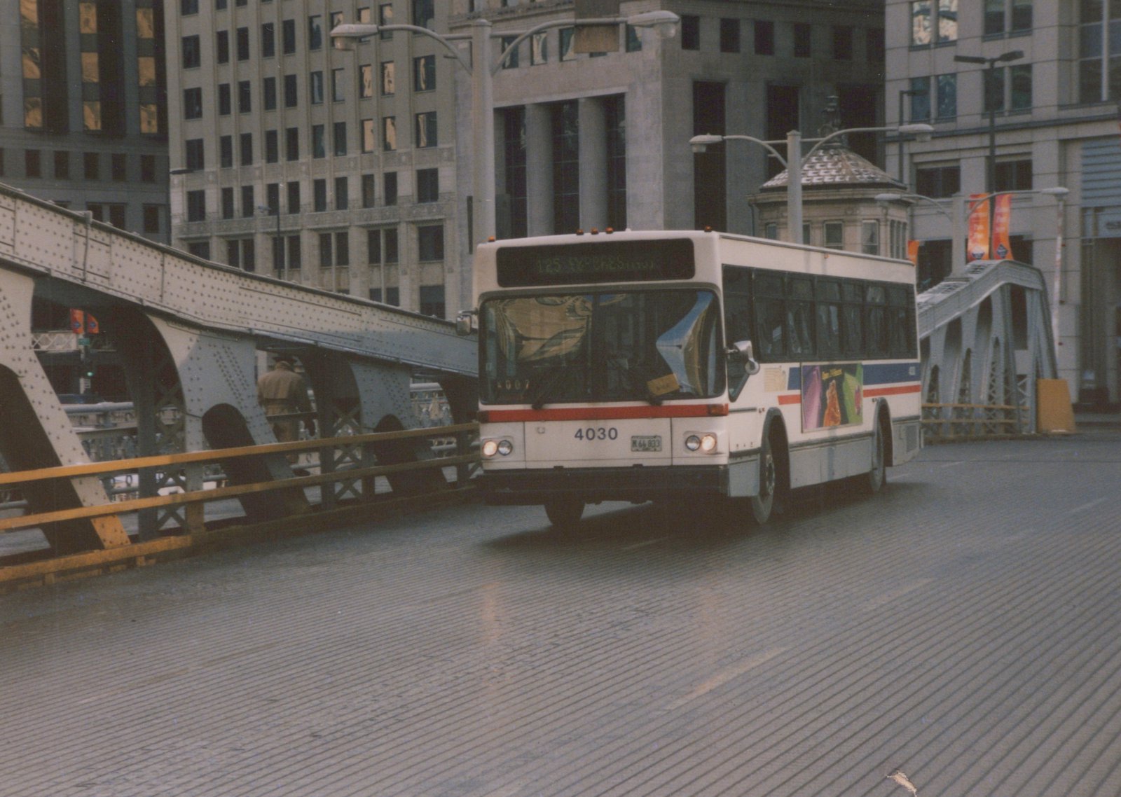 A white CTA bus, number 4030, is shown from a three-quarter front view driving on a street bridge in a city. A pedestrian walks on the bridge's walkway alongside the bus, with large buildings in the background.