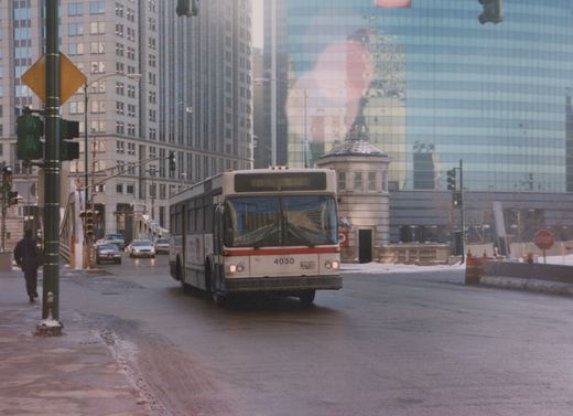 A white and red CTA bus with the number 4030 is driving on a wet, snow-dusted street in Chicago, with tall buildings in the background and a pedestrian on the sidewalk.