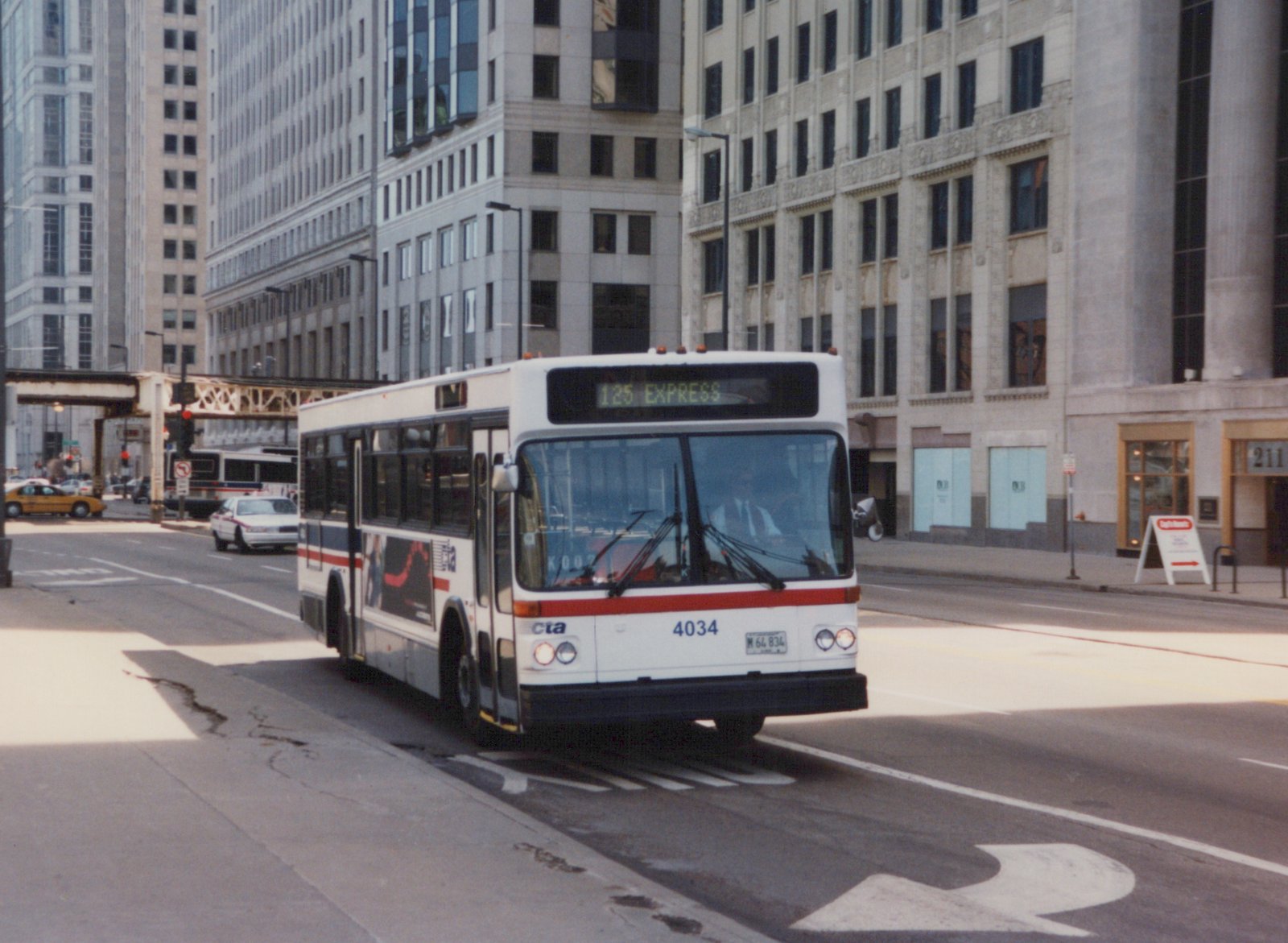 A white Chicago Transit Authority motor bus with red and blue stripes, displaying route 125 Express, drives on a city street with tall buildings in the background.